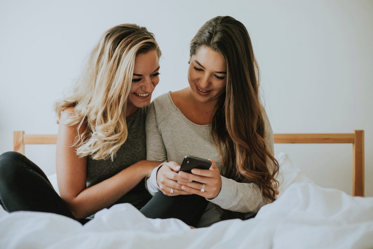 2 Frauen sitzen zusammen, lachen und schauen in ein Smartphone